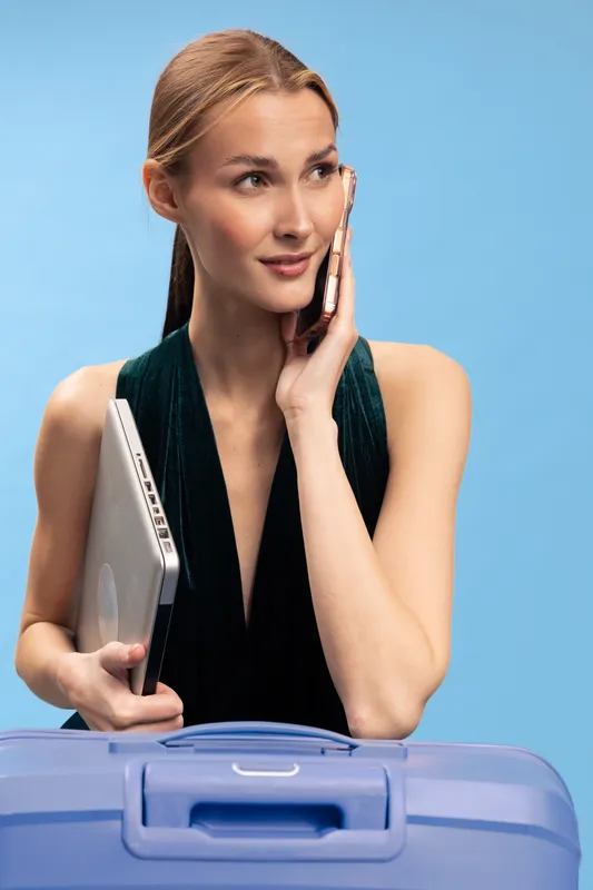 Young woman holds laptop and talks on smartphone near blue suitcase. She wears dark sleeveless dress and looks away on light blue background. Concept of travel and remote work.