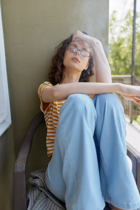 Casual woman relaxing on balcony, head resting on hand, sunlit afternoon, striped shirt and light blue pants, thoughtful mood, peaceful home lifestyle scene