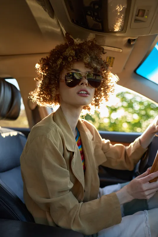 Young woman drives a car with retro style, curly hair, sunglasses, beige jacket, warm film color, casual lifestyle, daytime ride, cinematic mood, candid portrait inside a sunny vehicle