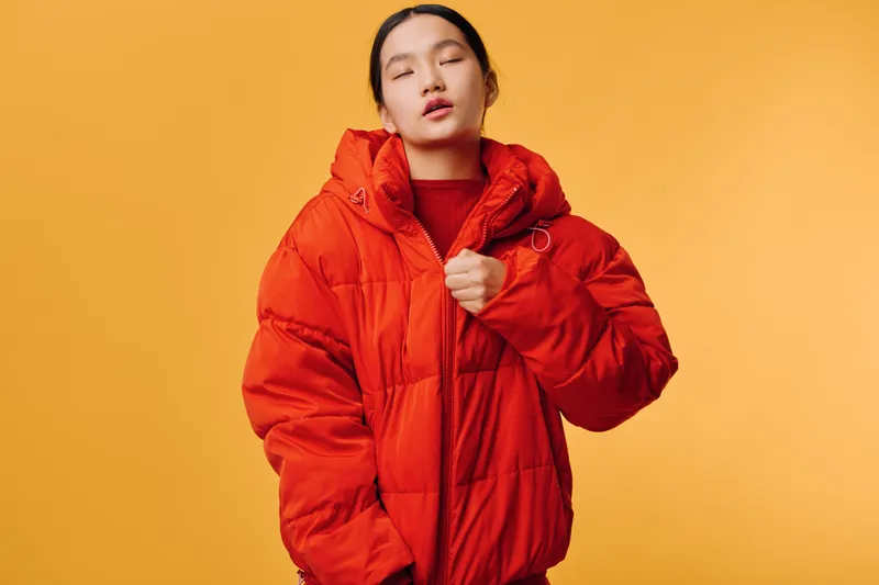 Young woman wearing bright red puffer jacket on yellow background, posing with closed eyes and relaxed expression. Casual fashion and winter clothing style portrait in studio.