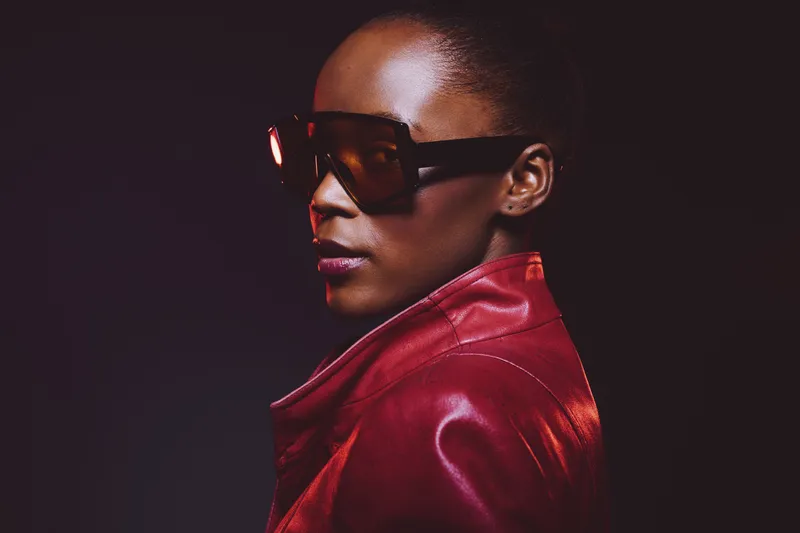 Portrait of confident black woman wearing large sunglasses and red leather jacket against dark background. Concept of fashion, style, elegance, and modern urban lifestyle.