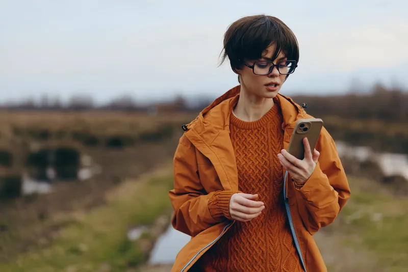 Woman lifestyle smartphone outdoors autumn portrait casual, orange jacket and sweater, holding smartphone while walking near countryside road, soft bokeh background, natural daylight, calm thoughtful
