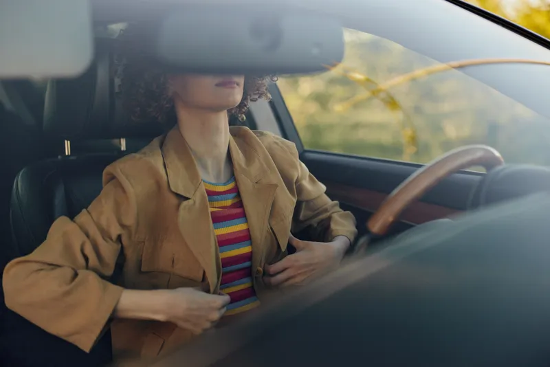 A woman driver sits in a car interior with retro vintage fashion, hands near chest, sunlight streaming through the window, warm tones and striped blazer evoke lifestyle calm.
