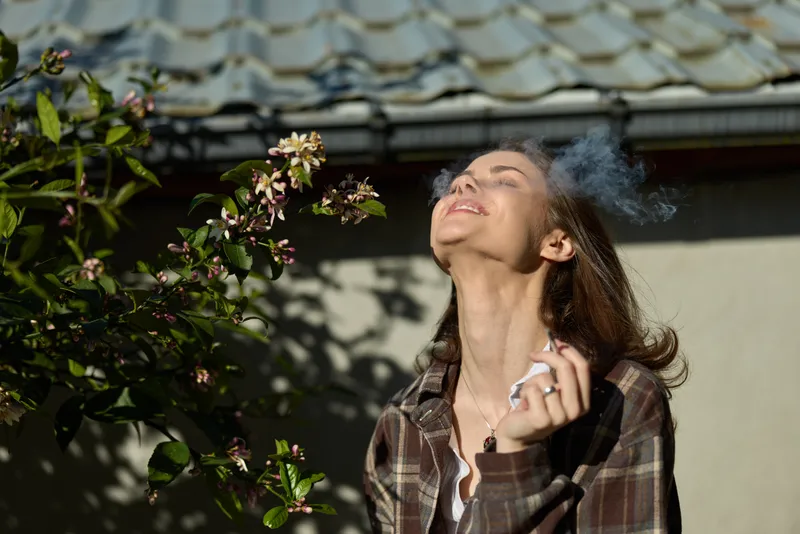 Joyful woman outdoors, smiling with head tilted up, sunlit moment, casual attire, breeze in hair, vibrant greenery backdrop, natural expression, relaxed lifestyle, candid moment