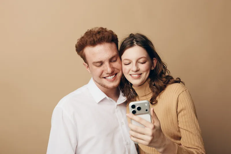 Smiling young couple with smartphone takes selfie together on beige background. They look happy and close, sharing a joyful moment of friendship and connection indoors.