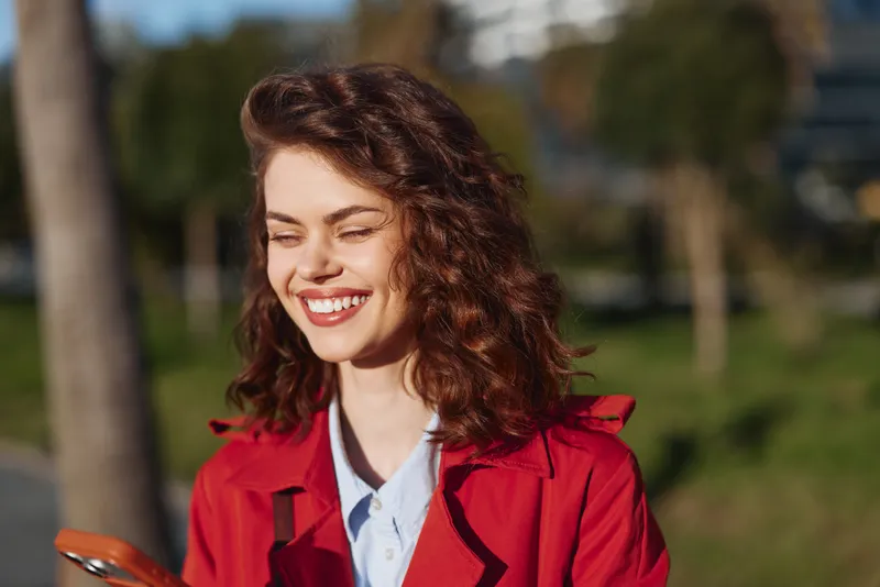 Young woman with curly hair in a red jacket smiles in an outdoor park on a sunny day, wearing a blue shirt and casual attire, exuding confidence and warmth while standing near green grass and trees,