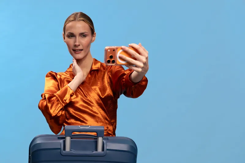 Young woman in orange satin blouse takes selfie with smartphone while holding suitcase handle on blue background. Travel concept with casual style and modern technology.