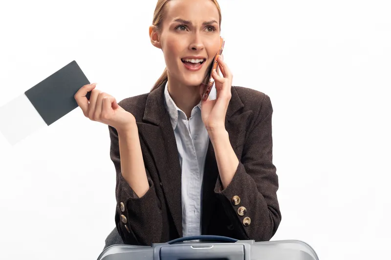 Young woman in business attire holds passport and talks on phone with excited expression. Concept of travel planning, business trip, and communication, with luggage in foreground on white background.
