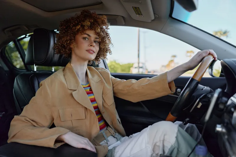 Smiling woman with curly hair sitting in a car wearing a beige jacket, lifestyle portrait with retro film color and warm daylight, relaxed urban mood. Ideal for lifestyle concepts, natural light and