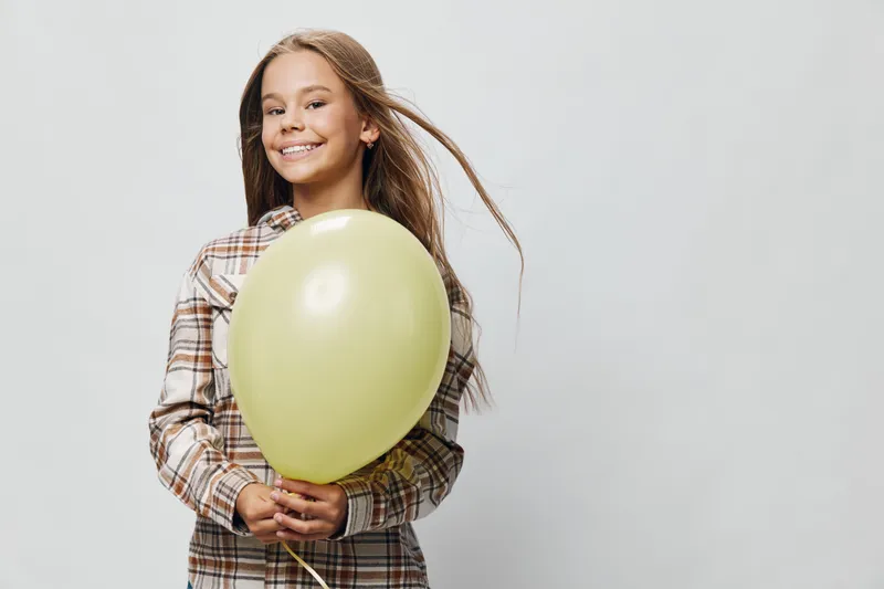 Girl smiling and holding yellow balloon wearing plaid shirt with long hair on light background. Child portrait with happy expression and casual clothing for celebration or party.