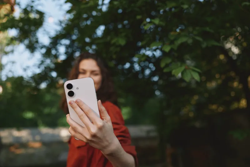 Cheerful woman outdoors, taking a selfie with a smartphone, casual red jacket, sunlit breeze, natural colors, relaxed posture, candid moment, lifestyle portrait outdoors ambiance