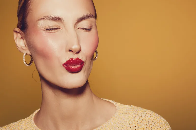 Close-up of a woman with red lipstick and gold hoop earrings making a kiss face. Warm yellow background and soft lighting create a stylish and playful mood.