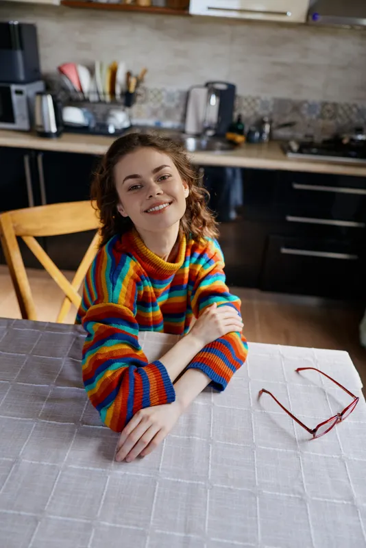 Smiling woman with curly hair wearing a colorful striped sweater sits at a kitchen table, arms rested on the surface. Cozy home setting with warm lighting conveys casual daily life.