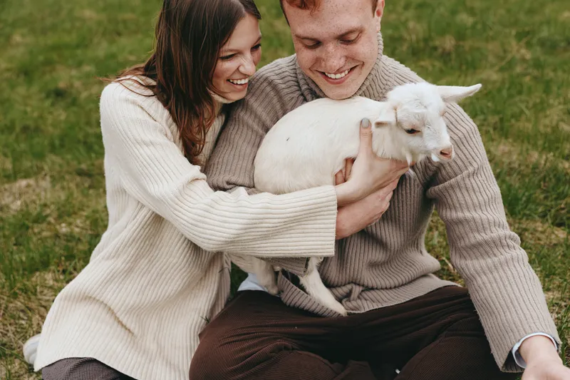 Young couple sitting on grass holding a white baby goat together while smiling and enjoying a relaxed moment in casual sweaters outdoors in a natural lifestyle scene.