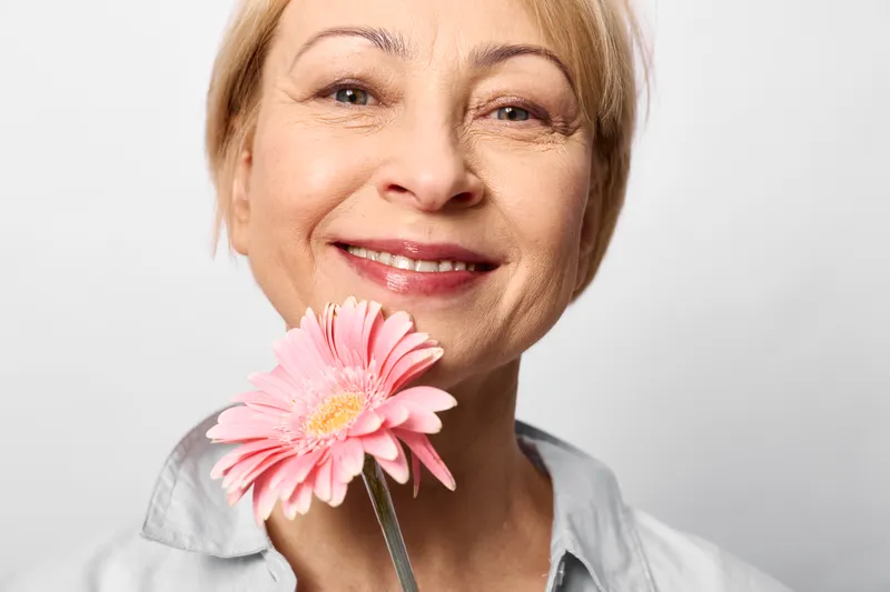 Smiling woman holding a pink flower in closeup elderly portrait, soft studio lighting, light background, calm happy mood, gentle expression, beauty, wellness, self care