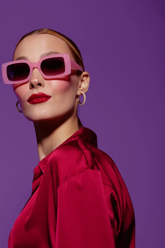 Stylish woman in pink sunglasses and red satin blouse poses confidently against purple background. Fashion portrait with bold makeup and modern accessories for trendy look.