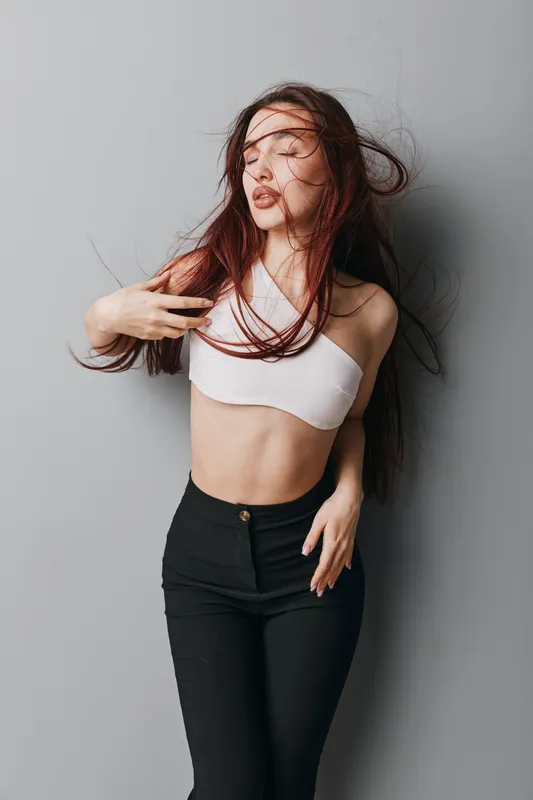 Young woman with long hair posing against gray wall wearing white crop top and black pants. Fashion model with closed eyes and hand in hair for beauty and style concept.