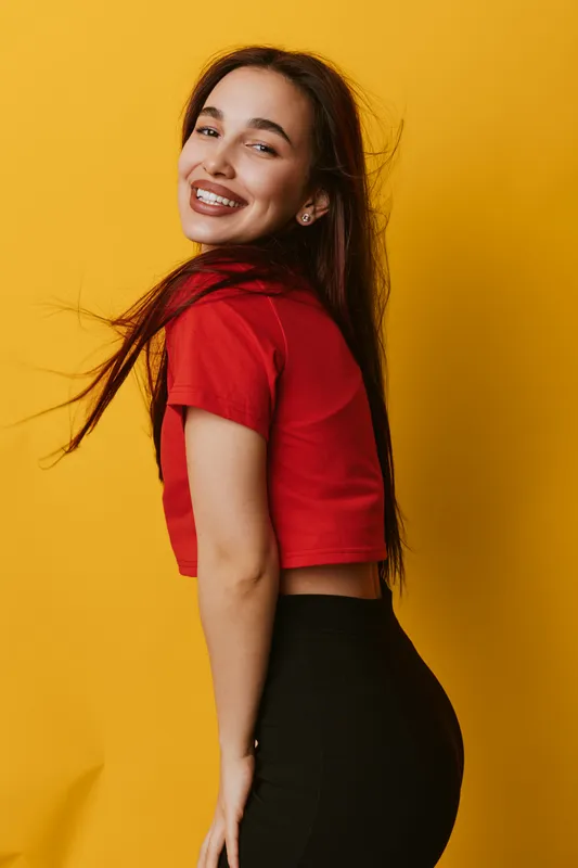 Young woman with long hair smiling and posing in red crop top and black pants against yellow background. Happy female model expressing joy and confidence in studio.