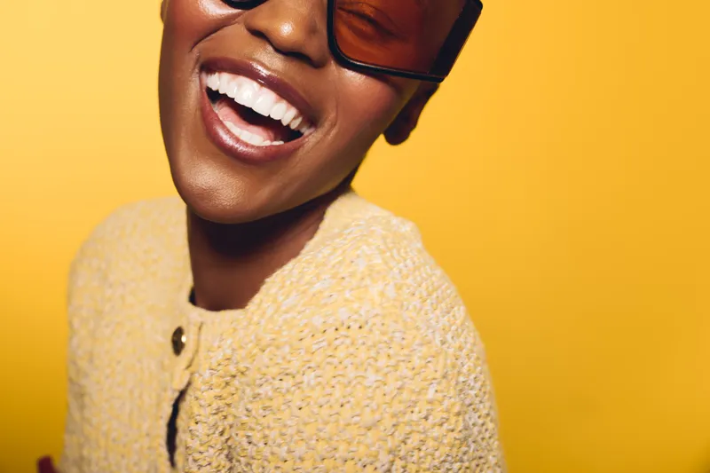 Happy African American woman with dark skin wearing sunglasses and a yellow knit sweater laughing against a bright yellow background. Concept of joy, fashion, and lifestyle.