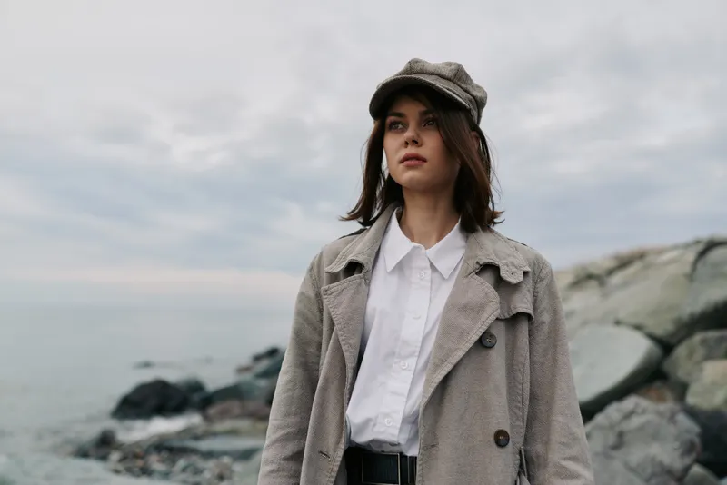 Young woman in stylish coat and hat stands near rocky shore under overcast sky. Fashion outfit and autumn mood captured in natural outdoor setting by the sea.