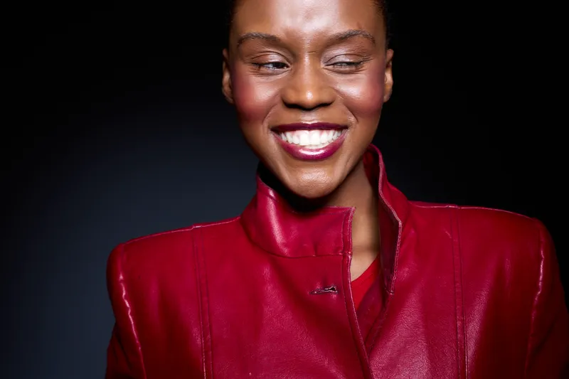 Smiling dark skinned African woman wearing bright red jacket winks with joy on black background. Concept of happiness, confidence, fashion, and positive mood in studio portrait.