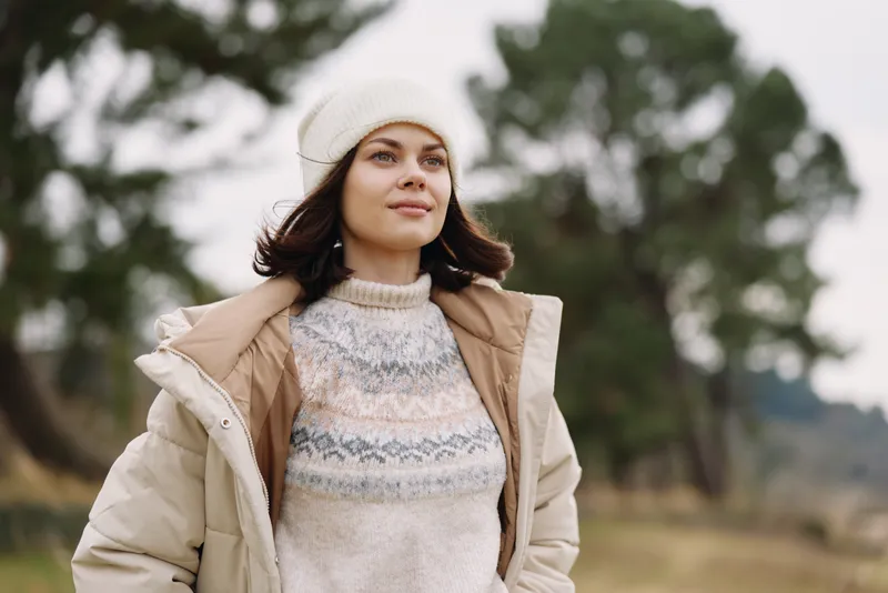 Woman standing outdoors in a warm coat and knit sweater, serene landscape behind, soft natural light, candid portrait capturing a calm winter mood during golden hour.