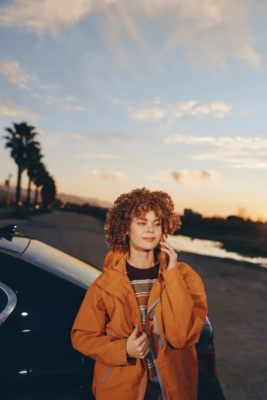 Woman lifestyle outdoors at sunset, curly hair and warm smile, wearing orange jacket, standing by car near beach road, coastal breeze, golden sunlight, relaxed travel vibe evening