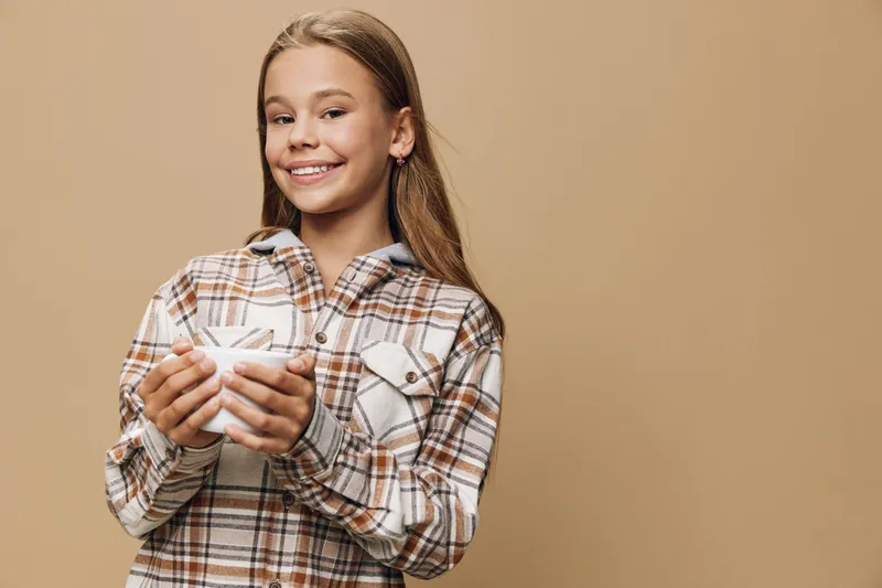 Girl smiling and holding a white cup standing against brown background. Casual clothes, long hair, plaid shirt, happy teenager with relaxed expression in studio portrait.