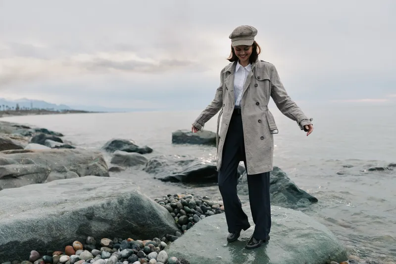 Young woman in elegant coat and hat walks on rocky beach by the sea. Overcast sky and calm water create a peaceful coastal atmosphere. Fashion and nature combined.