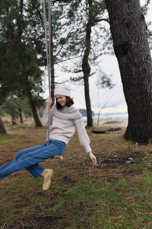 Woman on a rope swing in a park, outdoors among tall trees, wearing a knit sweater and jeans, enjoying a playful moment and fresh air in a peaceful nature setting.