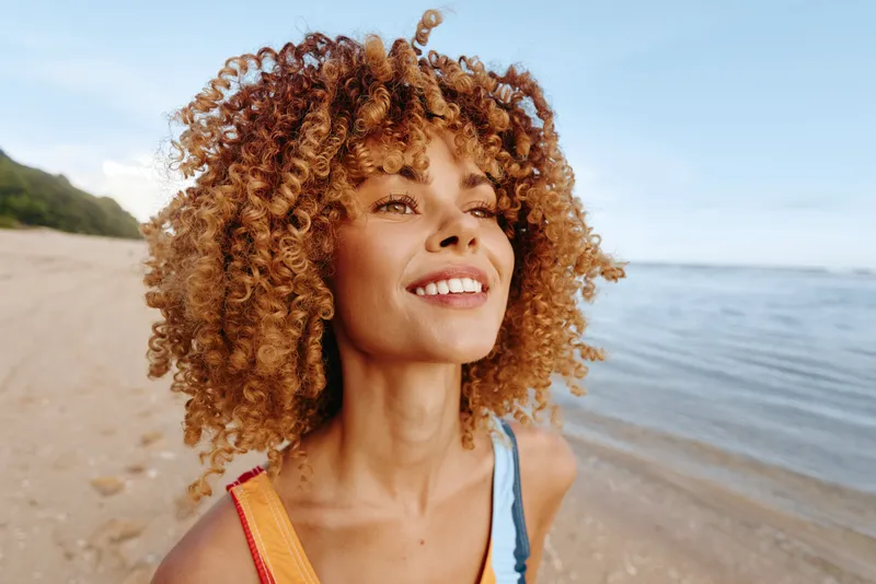 Bright portrait of a smiling woman on a sunny beach, relaxed pose, wind in curls, sparkling water behind, warm tones, soft sand, and natural mood conveying carefree summer joy
