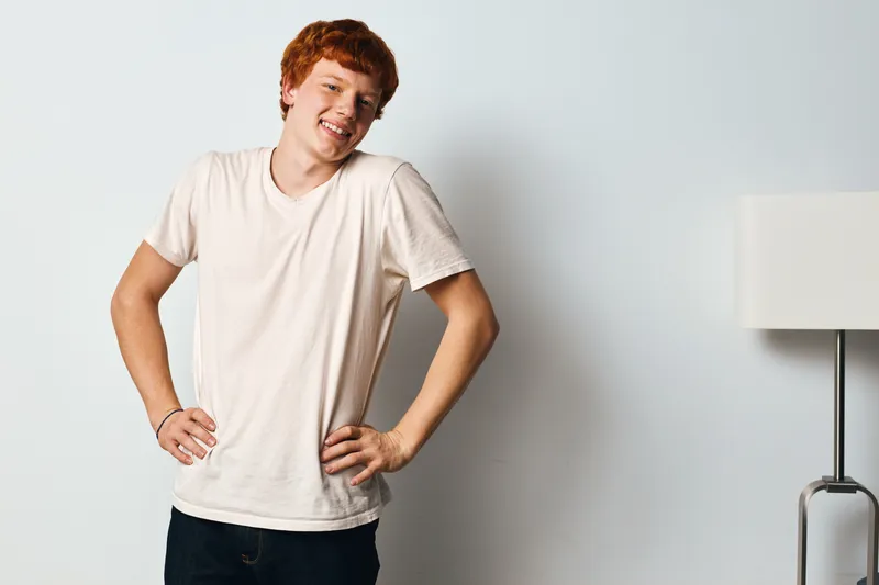 Teenager boy standing with hands on hips smiling confidently against white wall indoors. Casual young man with red hair and relaxed posture in simple light t shirt.