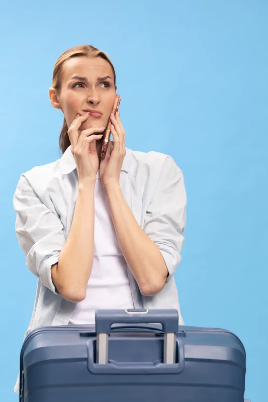 Young woman in white shirt stands with blue suitcase, holding phone to ear and looking thoughtful against blue background. Concept of travel planning, waiting, and communication.
