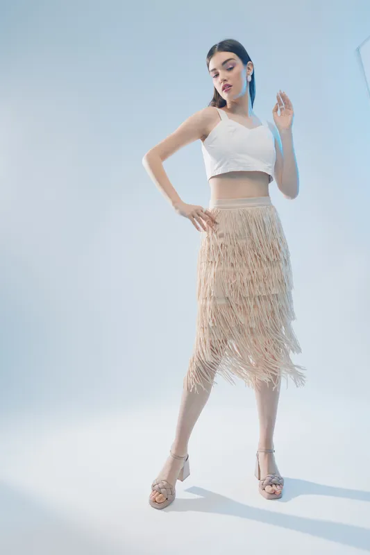 Young woman in a white crop top and beige fringe skirt poses confidently in a studio with soft lighting. Concept of beauty, fashion, and elegance.