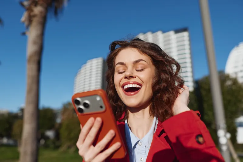 Young woman with curly hair in a red jacket laughs while looking at her smartphone outdoors, capturing a joyful moment in a sunny park with bright skies above the city skyline.