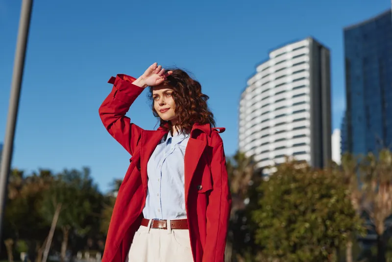 Cheerful woman outdoors in a red coat and white shirt, urban background with modern architecture, blue sky and greenery, sunlit casual street style in natural colors today