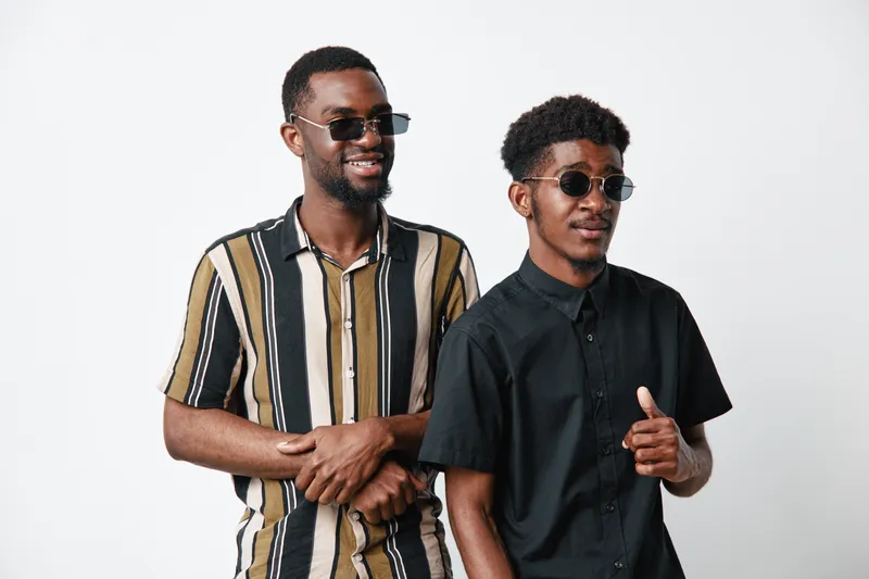 Two African American men with dark skin wearing sunglasses and casual shirts posing together in studio. Young friends smiling and showing thumbs up on white background.