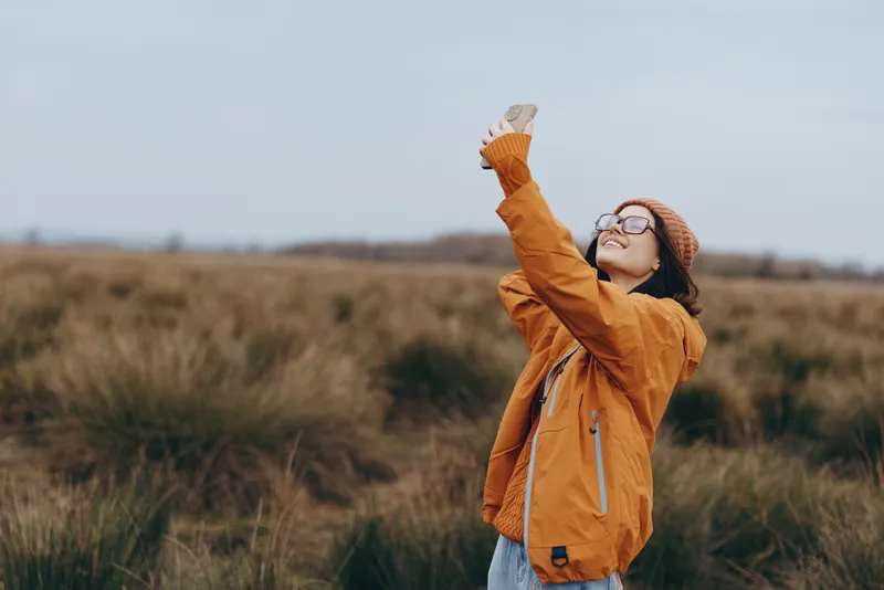 Smiling woman in outdoor autumn field, standing in nature landscape, wearing hat and scarf, lifting hands to adjust headwear, enjoying calm lifestyle, soft daylight and breeze
