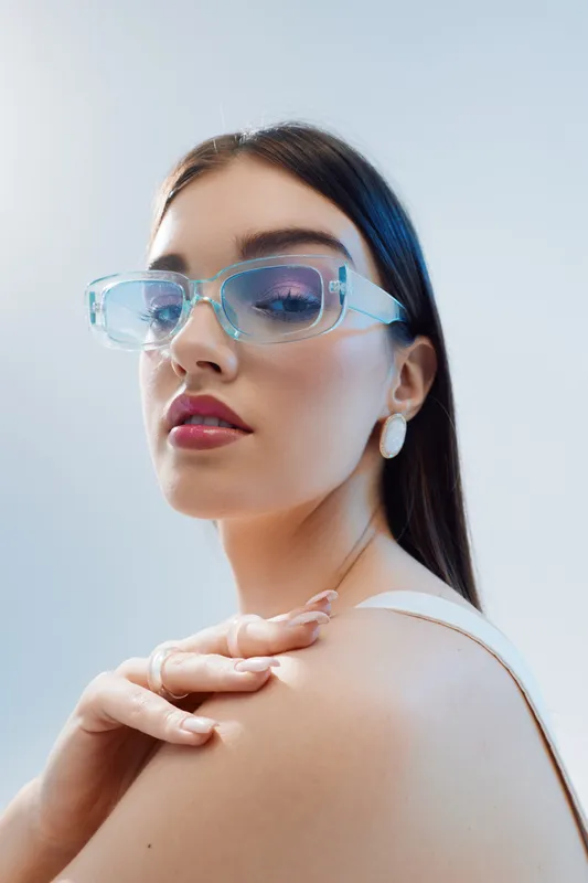 Young woman with long dark hair wearing transparent glasses and white earrings poses with hand on shoulder against a light background. Concept of beauty fashion and elegance.