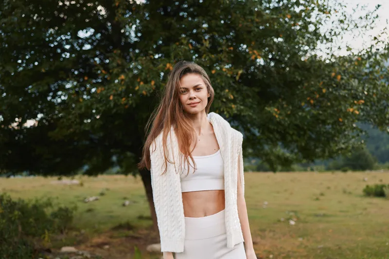 Young woman with long brown hair wears white knitted cardigan and crop top while standing outdoors in a natural green field with trees in the background.