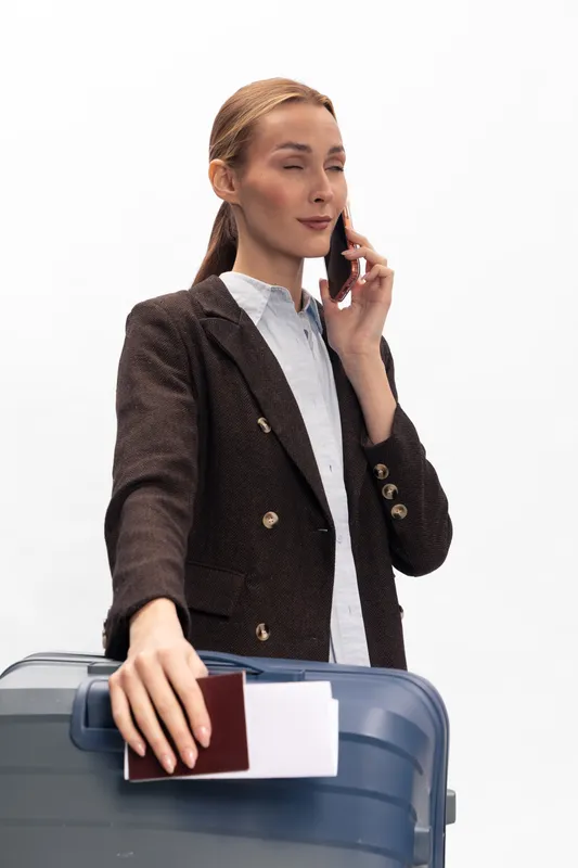 Businesswoman with suitcase holding passport and ticket talking on smartphone. Travel concept with professional woman preparing for trip in casual outfit isolated on white background.