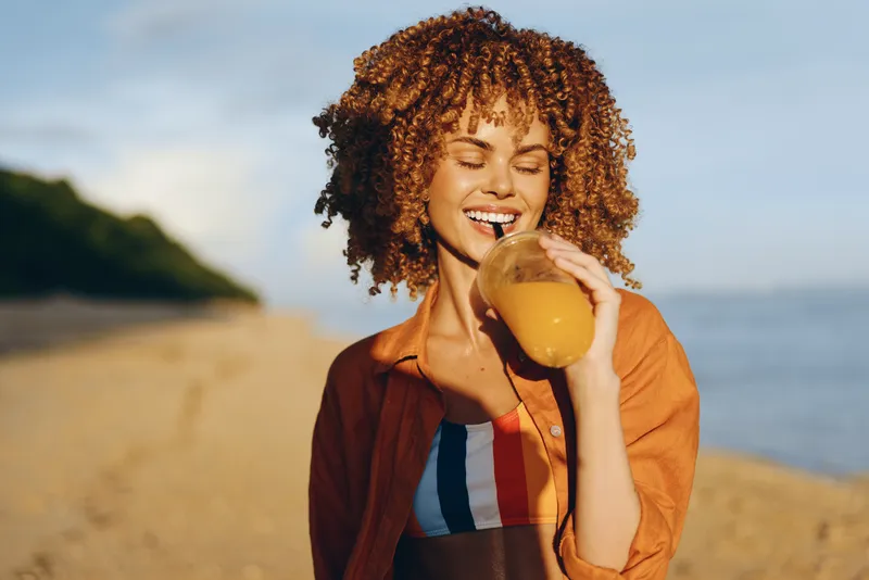 Woman with curly hair smiling while drinking orange juice on a sandy beach. Outdoor summer lifestyle with warm sunset light and relaxed wellness mood. Casual maroon jacket, striped top, candid moment.