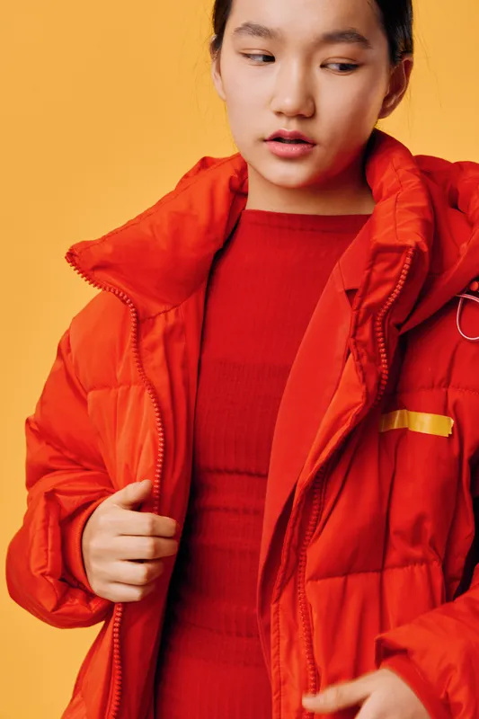 Young woman wearing a bright red winter jacket over red dress posing against a solid orange background. Fashion model with casual look in warm clothing for cold weather style portrait.