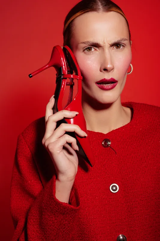 Woman in red sweater holds red high heel shoe near face with confused expression on red background. Concept of fashion, style, emotions, and creative portrait photography.