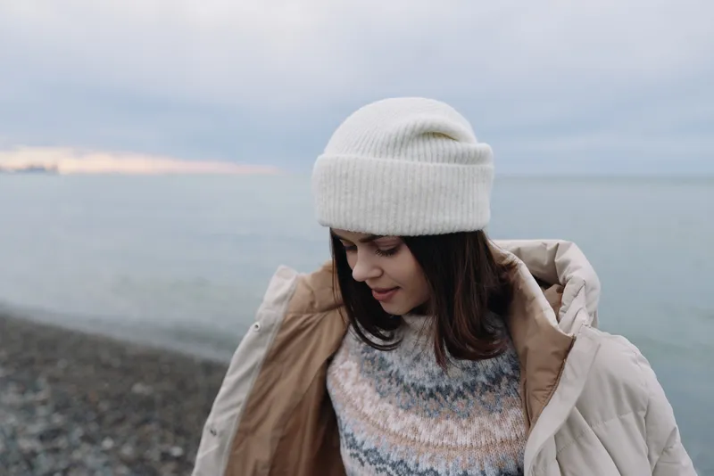 Woman in a knit sweater and warm coat on a windy beach, winter landscape by the sea, soft light and calm mood.