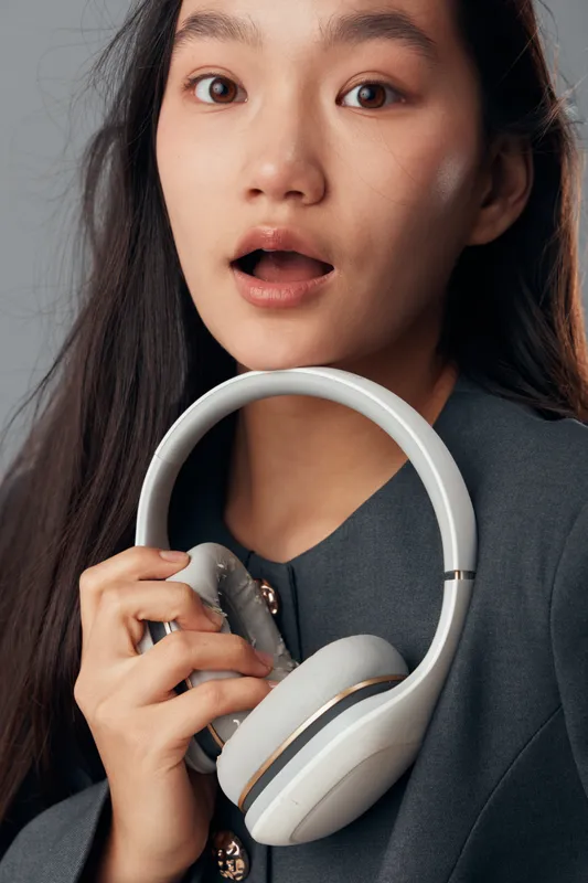 Woman with surprised expression holding white headphones close to her face. Portrait of female in casual dark clothing with long hair on grey background with open mouth and wide eyes.