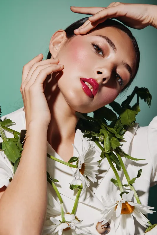 Creative beauty portrait of a young woman wearing bold makeup and surrounded by fresh flowers on a green background. Artistic concept for fashion and cosmetics.