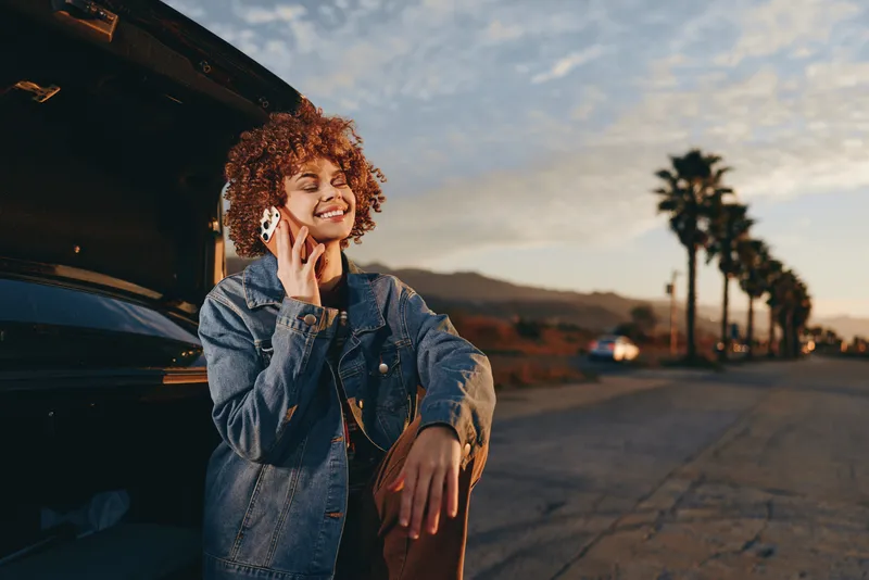 Smiling woman lifestyle outdoors near car in warm sunset light, curly hair, denim jacket, relaxed pose, playful phone gesture, open road background, soft breeze, natural colors evening