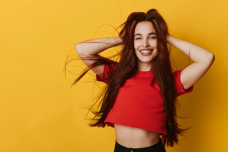 Young woman with long hair wearing red crop top smiling and holding hands behind head against yellow background. Casual style, happiness and carefree mood in studio portrait.