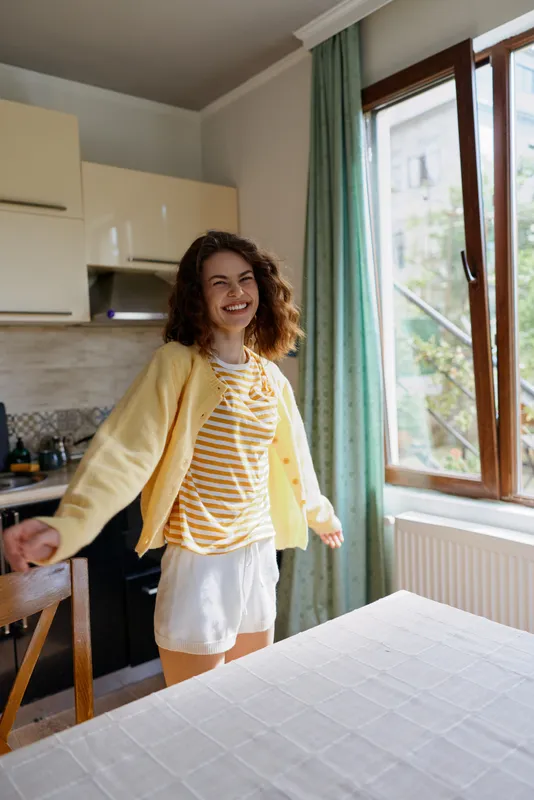 Young woman with curly hair smiles and spins in a bright kitchen, wearing a striped shirt and yellow cardigan, enjoying a relaxed home moment by the sunlit window